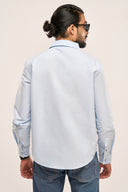 Close up back view of a man wearing a light blue chambray shirt with apple-cut hem and full sleeves, paired with jeans.