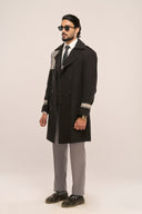 Man in black trench coat with plaid accents, waist belt, paired with grey pants, black shoes, and sunglasses, side view.