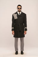Man in black trench coat featuring plaid accents, waist belt, horn buttons, paired with grey pants, black shoes & sunglasses.