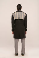 Rear view of man in black trench coat with plaid shoulder overlay, gray pants, standing against plain background.