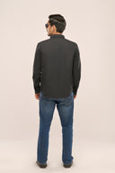 Man seen from behind wearing a charcoal black chambray shirt with full sleeves and apple-cut hem, paired with blue jeans.