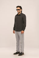 Man wearing a slim-fit charcoal black pleat shirt with full sleeves, light pants, and black shoes on a beige background.